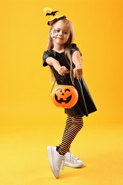 Photo of Cute girl with pumpkin bucket dressed like witch on yellow background. Halloween celebration