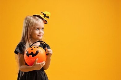 Photo of Cute girl with pumpkin bucket dressed like witch on yellow background, space for text. Halloween celebration