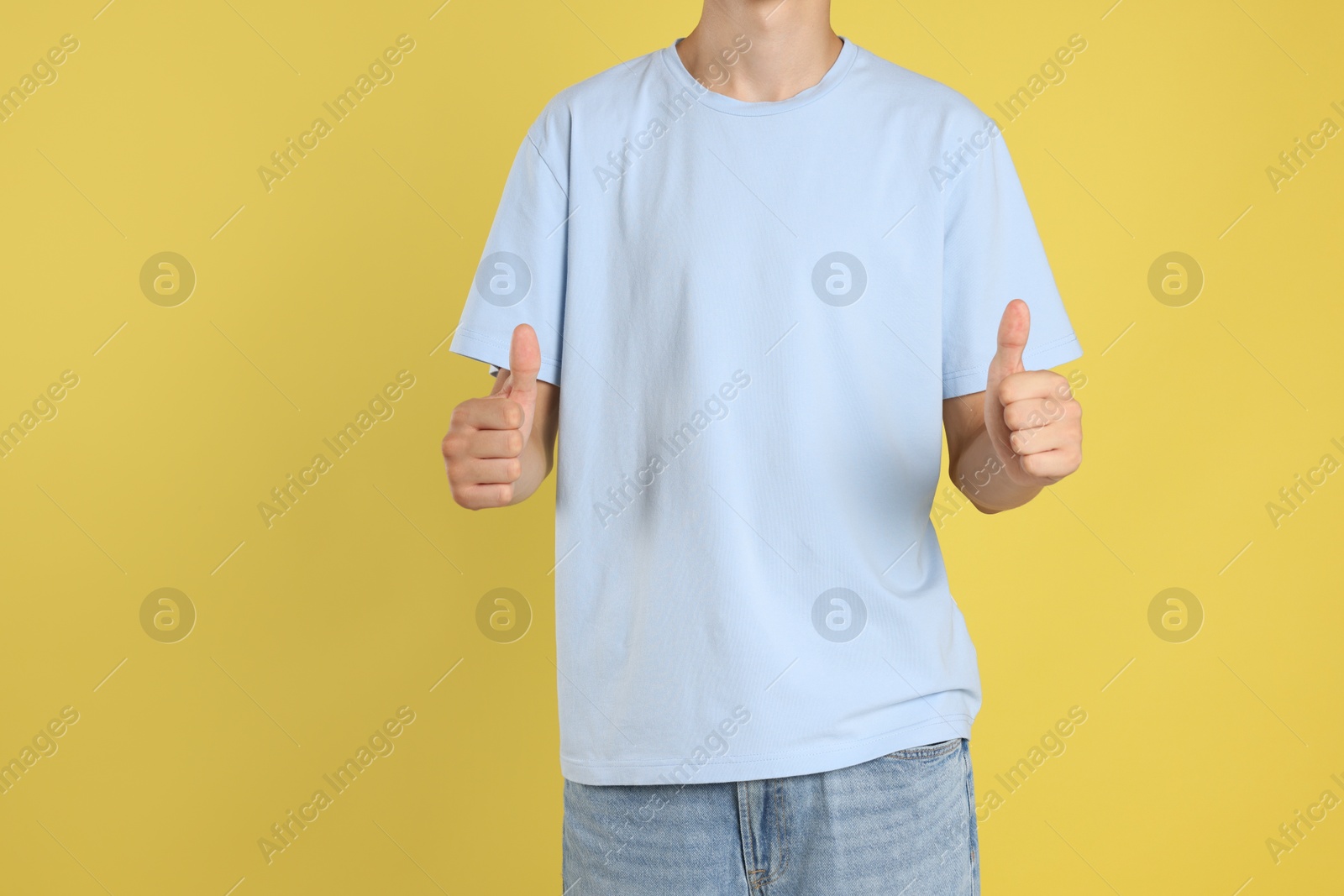 Photo of Teenage boy wearing light blue t-shirt and showing thumbs up on yellow background, closeup. Space for text