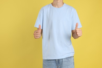 Photo of Teenage boy wearing light blue t-shirt and showing thumbs up on yellow background, closeup. Space for text