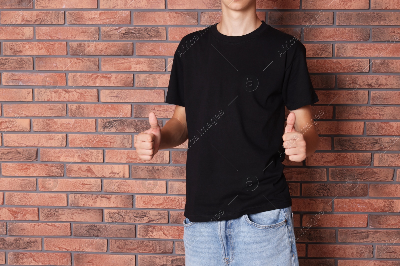 Photo of Teenage boy wearing black t-shirt and showing thumbs up near brick wall, closeup. Space for text