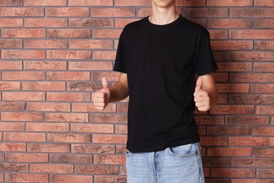 Photo of Teenage boy wearing black t-shirt and showing thumbs up near brick wall, closeup. Space for text