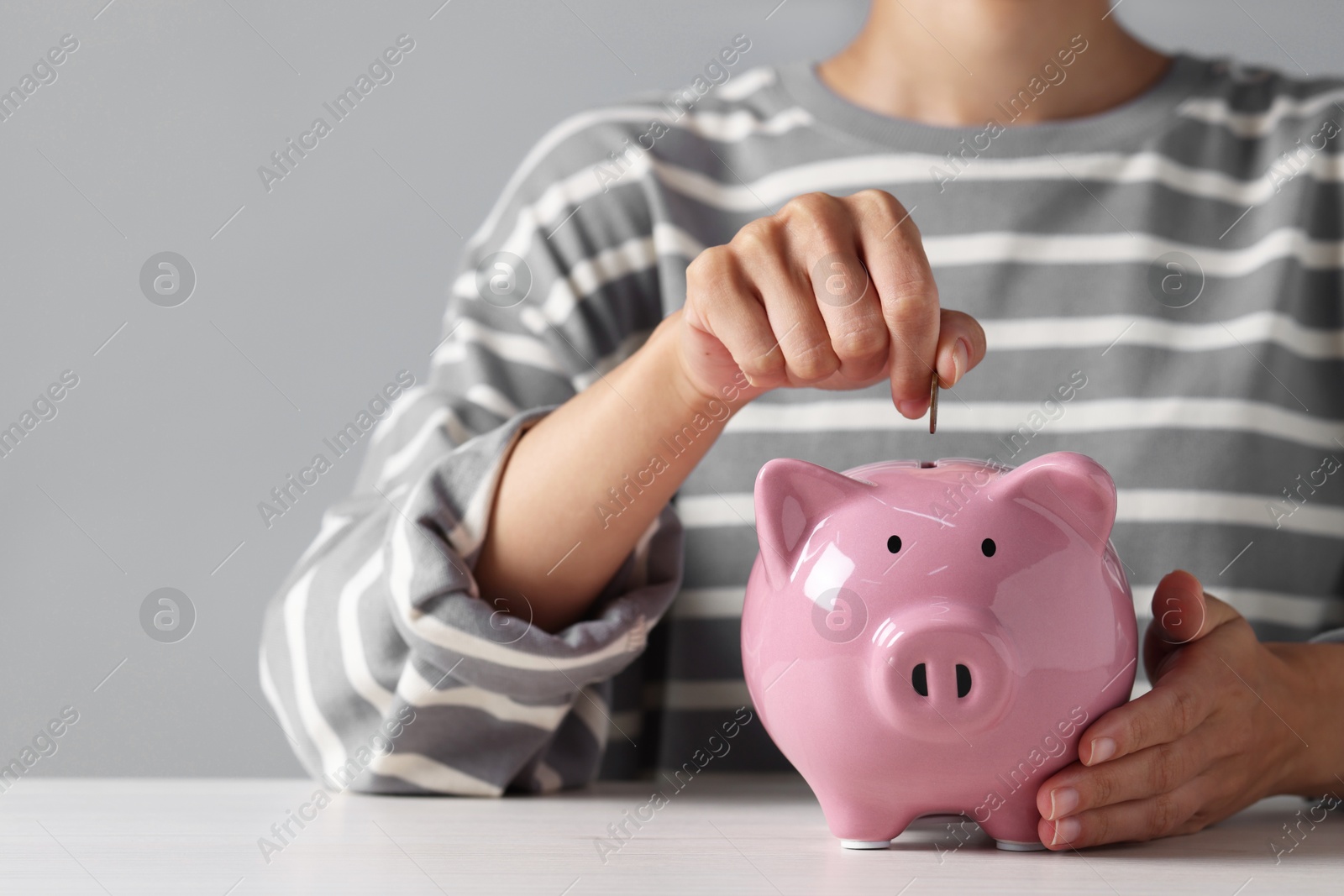 Photo of Woman putting coin into piggy bank at white wooden table, closeup