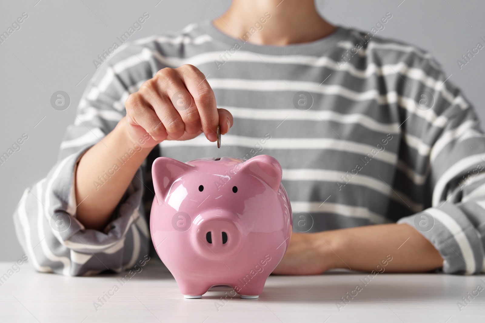 Photo of Woman putting coin into piggy bank at white wooden table, closeup