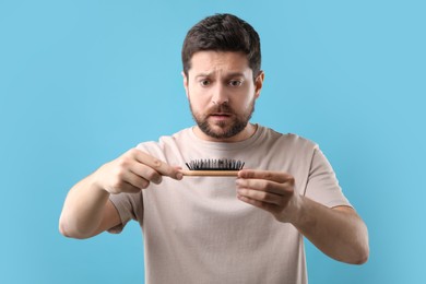 Photo of Sad man holding brush with lost hair on light blue background. Alopecia problem