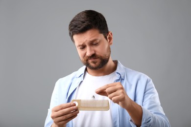 Sad man taking his lost hair from comb on gray background. Alopecia problem