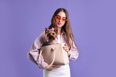 Beautiful young woman carrying cute Yorkshire Terrier dog in bag on violet background