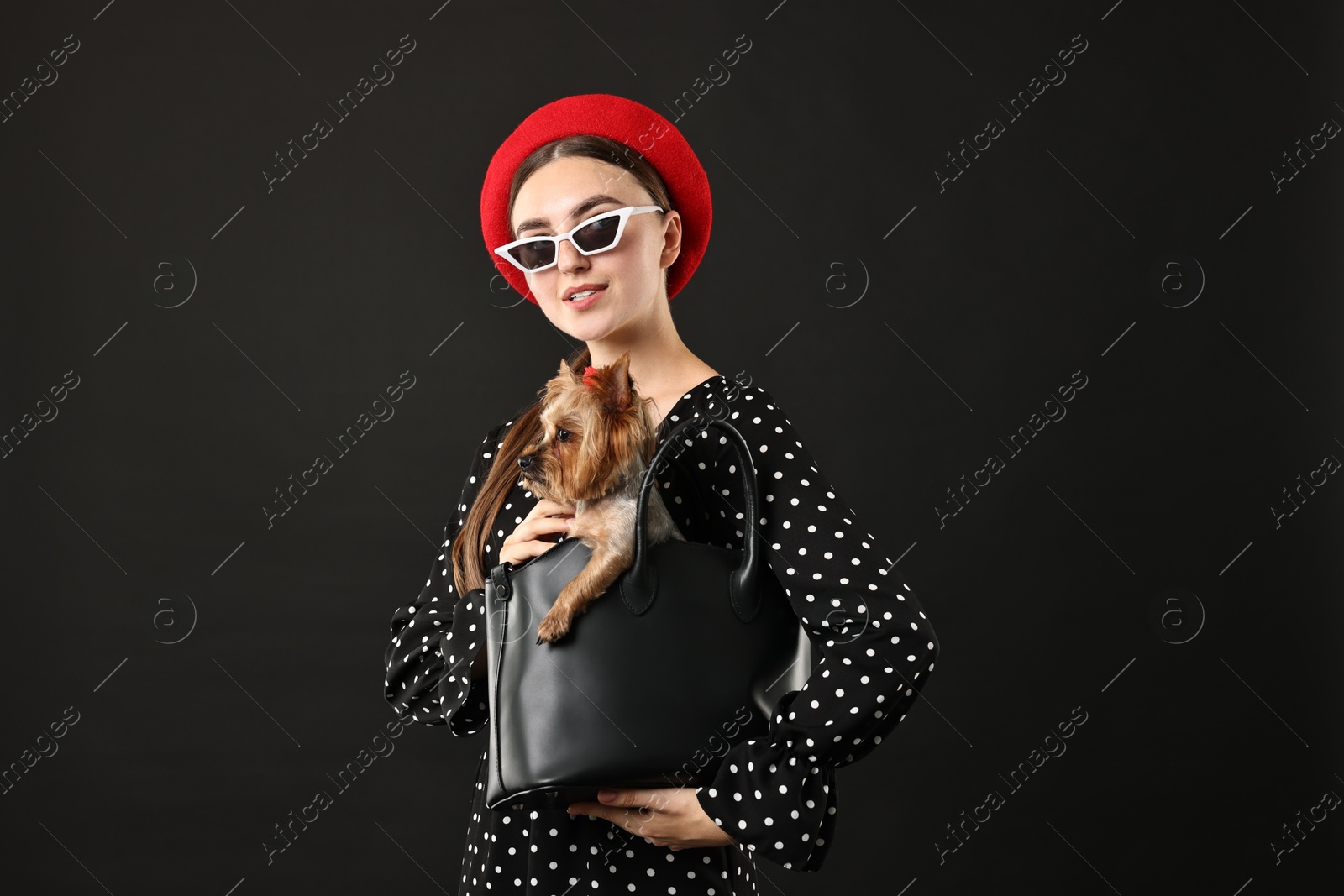 Photo of Woman in sunglasses holding bag with cute Yorkshire Terrier dog on black background