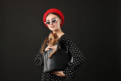 Woman in sunglasses holding bag with cute Yorkshire Terrier dog on black background