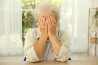 Senior woman closing her eyes with hands at wooden table indoors