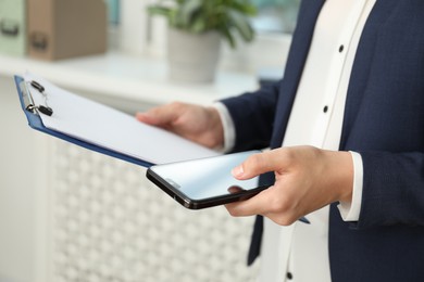 Businesswoman with clipboard using smartphone indoors, closeup. Modern technology