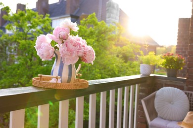 Photo of Vase with beautiful peonies on railings and chair at cozy balcony on sunny day