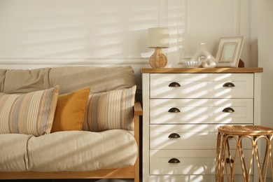 Photo of Sofa with soft pillows, chest of drawers, different decorative elements and side table indoors