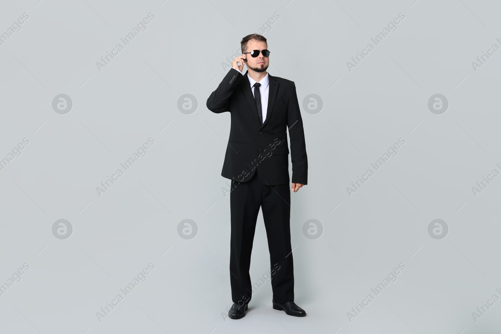 Photo of Young bodyguard in suit and sunglasses using earpiece on grey background