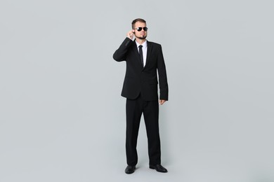 Young bodyguard in suit and sunglasses using earpiece on grey background