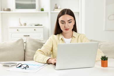 Photo of Budget planning. Young woman with papers using laptop at white table indoors