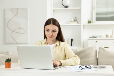 Budget planning. Young woman with papers using laptop at white table indoors