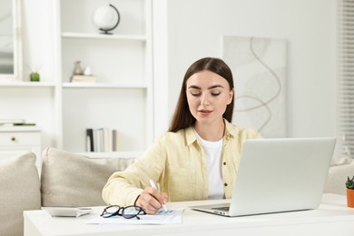 Photo of Budget planning. Young woman with papers using laptop at white table indoors