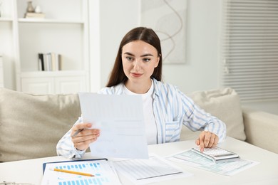 Budget planning. Beautiful young woman with papers using calculator at white table indoors