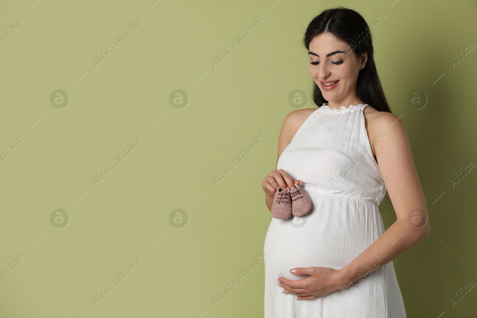 Photo of Portrait of beautiful pregnant woman with baby shoes on light olive background, space for text