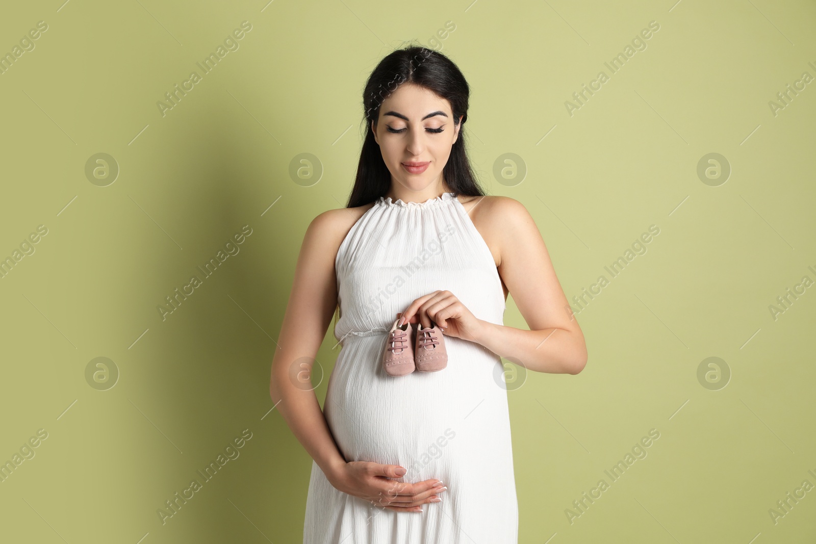 Photo of Portrait of beautiful pregnant woman with baby shoes on light olive background