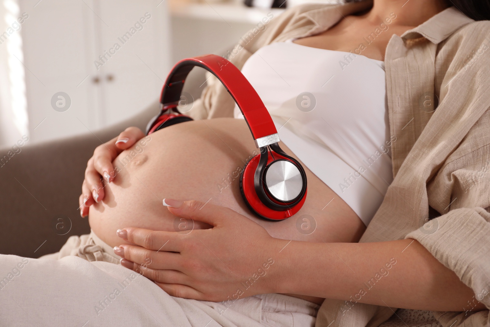 Photo of Young pregnant woman with headphones on belly at home, closeup