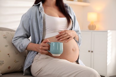 Photo of Young pregnant woman with cup of drink sitting on sofa at home, closeup