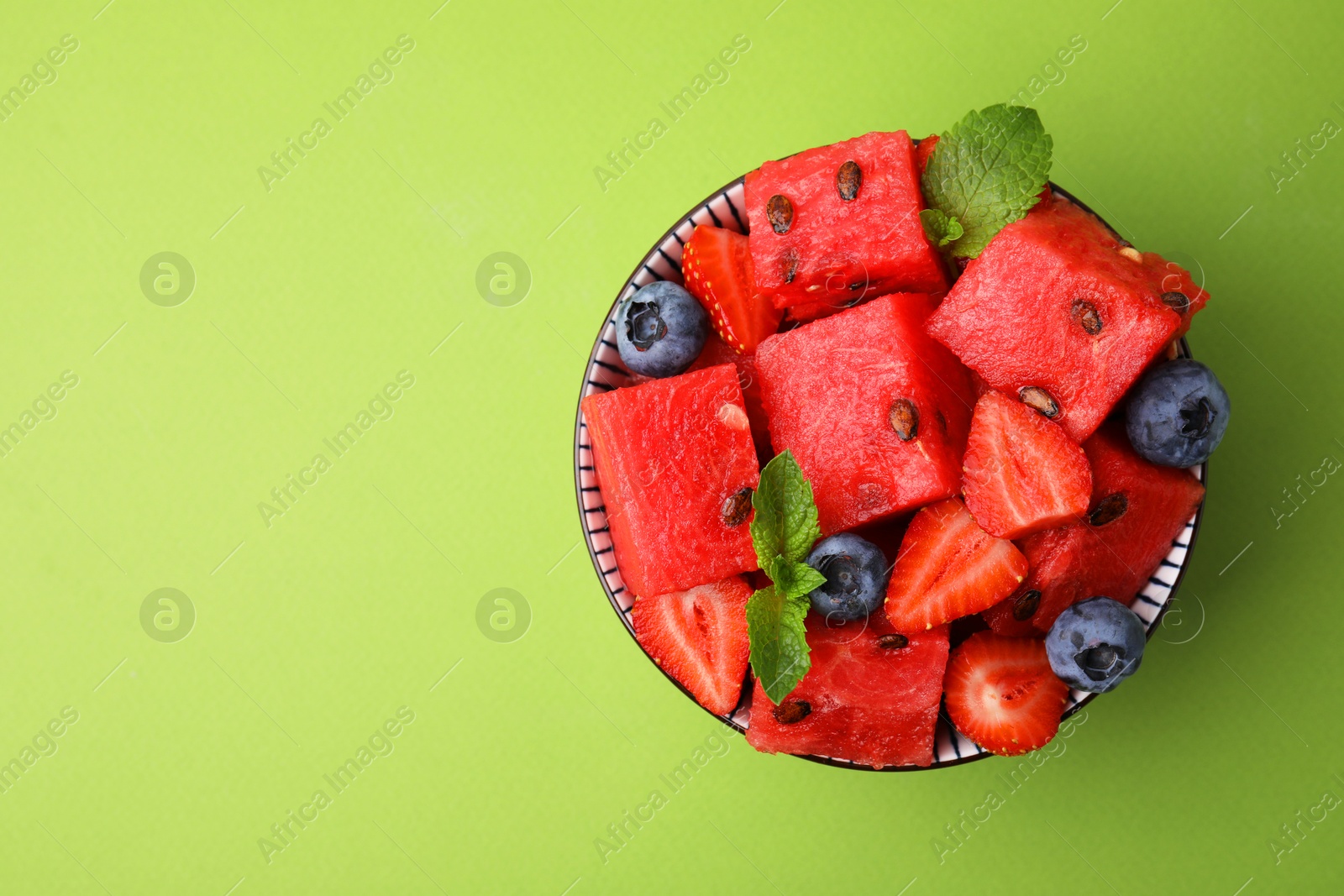 Photo of Pieces of tasty watermelon, strawberries, blueberries and mint in bowl on green table, top view. Space for text