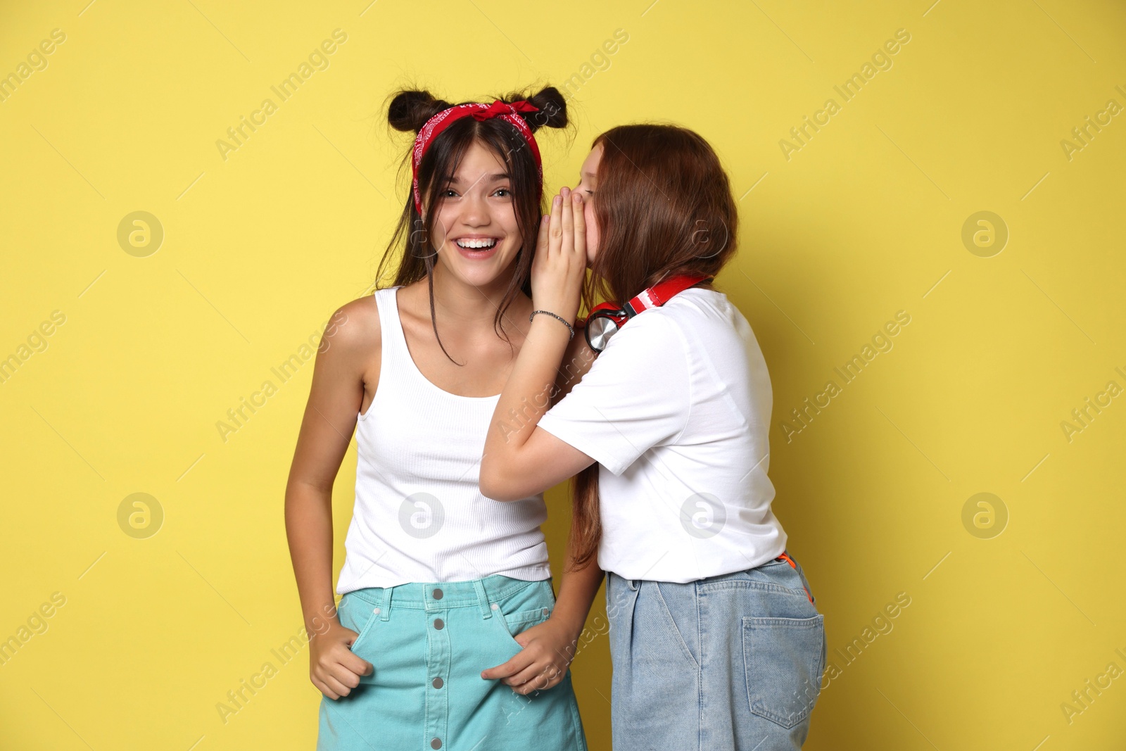 Photo of Teenage girl whispering secret to her happy friend on yellow background