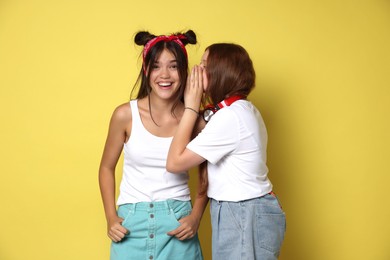 Teenage girl whispering secret to her happy friend on yellow background