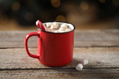 Photo of Tasty hot cocoa drink with candy cane and marshmallows in red mug on wooden table, closeup