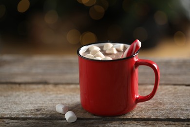 Photo of Tasty hot cocoa drink with candy cane and marshmallows in red mug on wooden table, closeup