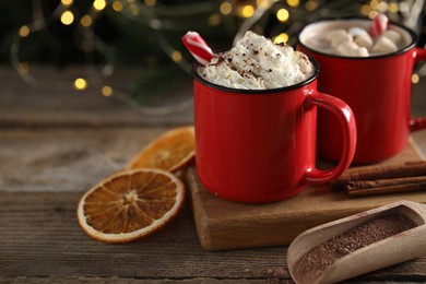 Photo of Tasty hot cocoa drink with whipped cream in red mugs and dry orange on wooden table