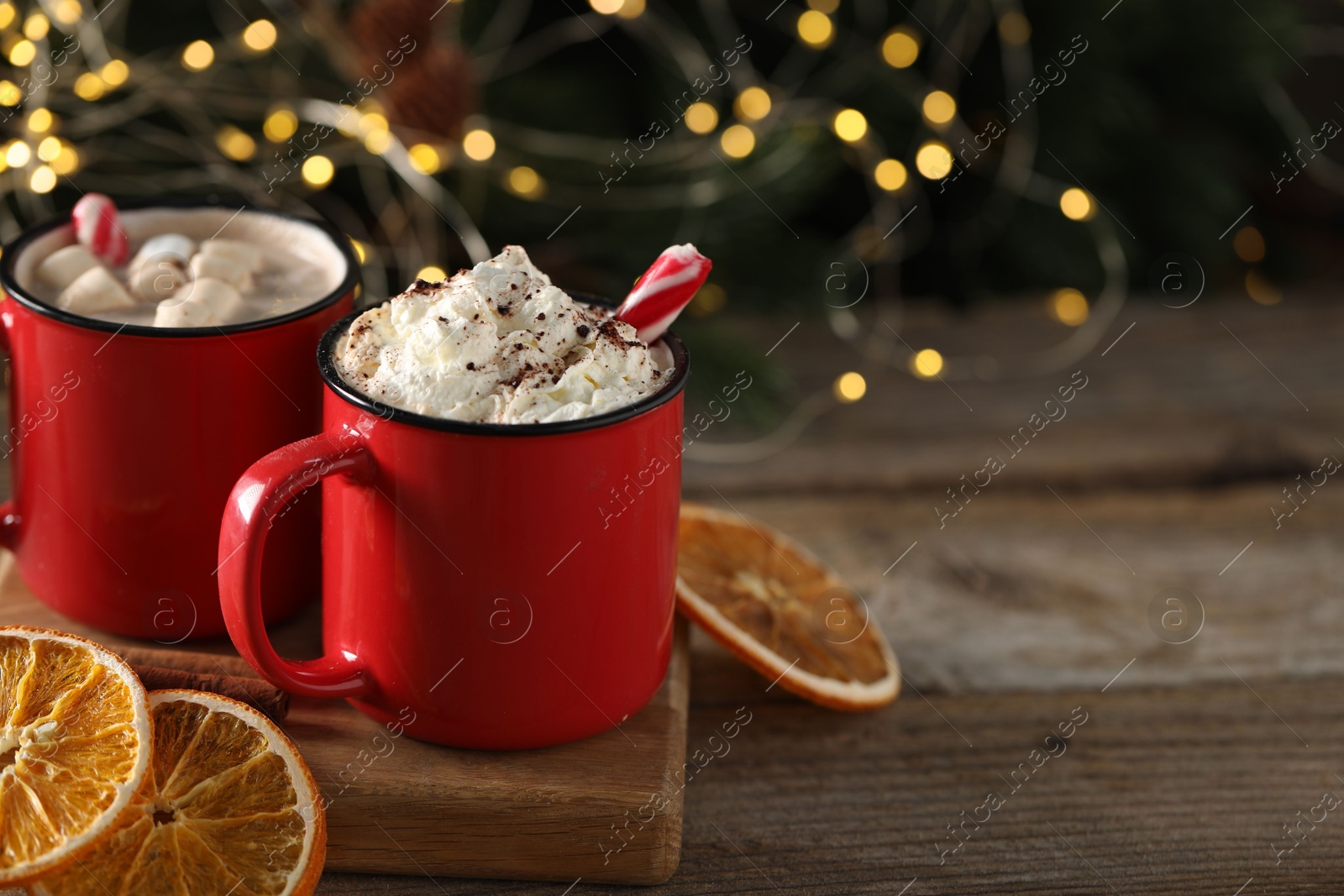 Photo of Tasty hot cocoa drink with whipped cream in red mugs and dry orange on wooden table, space for text