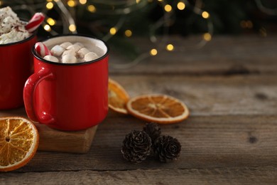 Photo of Tasty hot cocoa drink with marshmallows in red mugs, dry orange and cones on wooden table, space for text