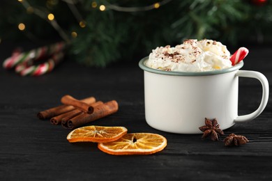 Photo of Tasty hot cocoa drink with whipped cream in mug and spices on black wooden table, closeup