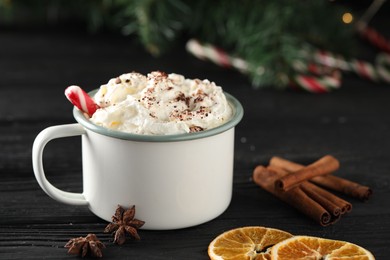 Photo of Tasty hot cocoa drink with whipped cream in mug and spices on black wooden table, closeup