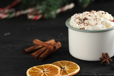 Photo of Tasty hot cocoa drink with whipped cream in mug and spices on black wooden table, closeup. Space for text