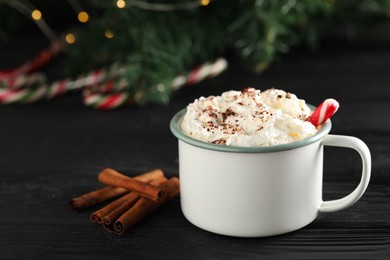 Photo of Tasty hot cocoa drink with whipped cream in mug and spices on black wooden table, closeup