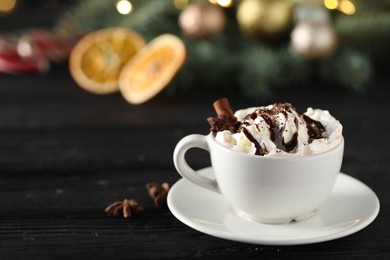 Photo of Tasty hot cocoa drink with whipped cream in cup and spices on black wooden table, space for text