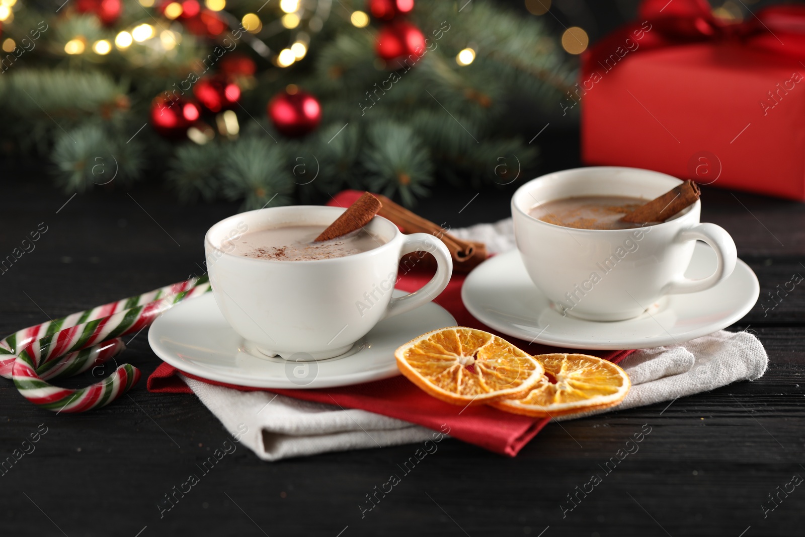 Photo of Tasty hot cocoa drink with cinnamon sticks in cups, dry orange and candy canes on black wooden table