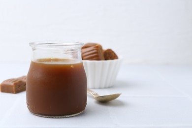 Photo of Tasty caramel sauce in jar, spoon and candies on white tiled table, closeup. Space for text