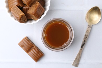 Photo of Tasty caramel sauce in jar, spoon and candies on white tiled table, flat lay