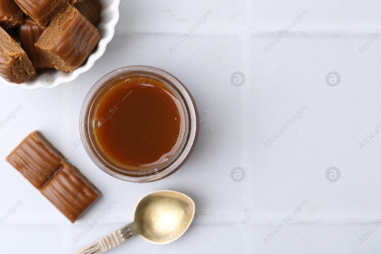 Photo of Tasty caramel sauce in jar, spoon and candies on white tiled table, flat lay. Space for text