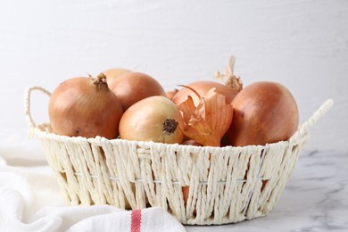 Fresh onions with peels in wicker basket on light marble table