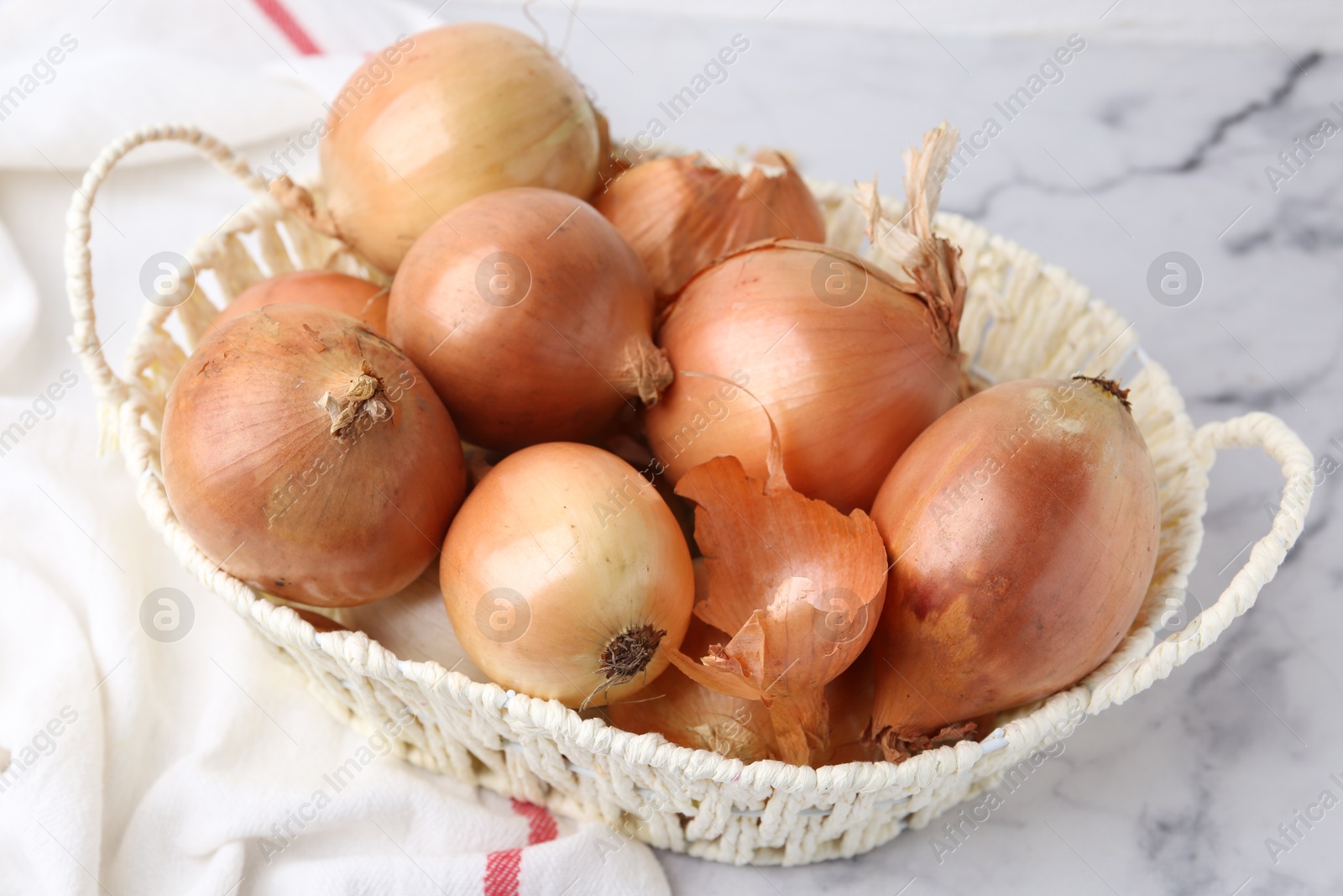 Photo of Fresh onions with peels in wicker basket on light marble table, closeup