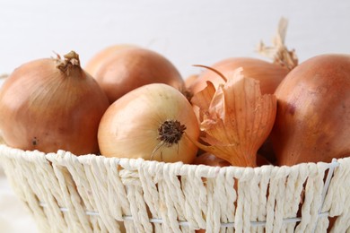 Fresh onions with peels in wicker basket against light background, closeup