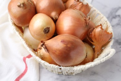 Photo of Fresh onions with peels in wicker basket on light marble table, closeup