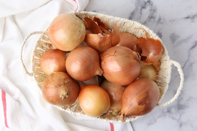 Photo of Fresh onions with peels in wicker basket on light marble table, top view