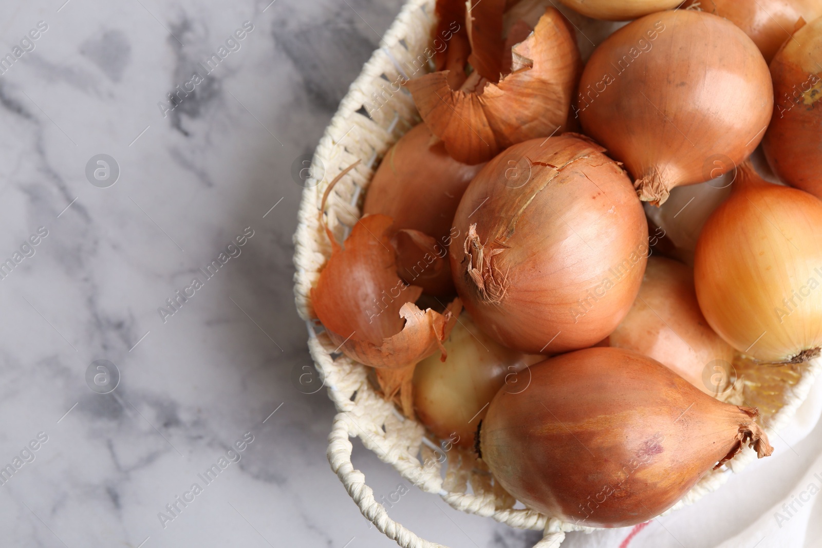 Photo of Fresh onions with peels in wicker basket on light marble table, top view. Space for text